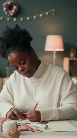 Vertical shot of focused African American woman writing warm wishes on Christmas card while sitting at cluttered with colorful pencils table in decorated living room