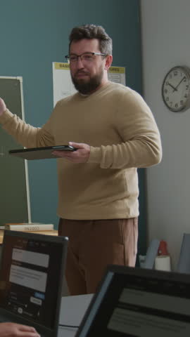 Medium vertical shot of cheerful Caucasian male information technology teacher explaining AI generator prompt algorithm to offscreen students, working on laptops in classroom