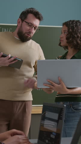 Medium vertical shot of young Caucasian female student asking cheerful male IT teacher for advice on coding project, mentor checking and giving feedback, suggesting corrections, pointing to mistakes