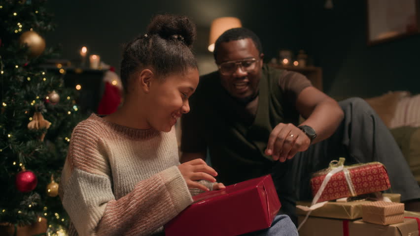 Joyful Black girl child opening wrapped gift box receiving Christmas present from loving father while sitting on floor in decorated living room in evening