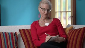 A happy mature woman relaxes on her couch writing in journal, wearing a heart sensor to monitor atrial fibrillation and improve her health. - Powered by Shutterstock - Get 15% off with code: PIKWIZARD15