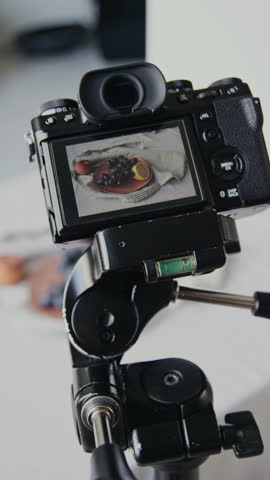Vertical shot of professional camera on black tripod, screen demonstrating plate with black grape and lemon