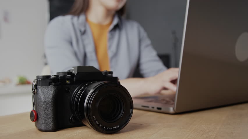 Close up of professional black camera on wooden desk, unrecognizable woman typing on keyboard of silver laptop in blurred background