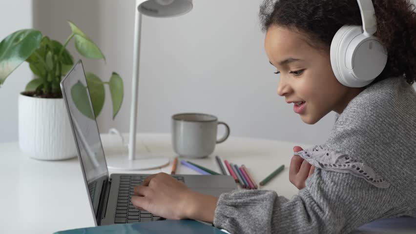Primary school African American Student child with headphones hand writing in book using laptop. Distance learning online education. School girl studying at home with digital tablet computer and doing