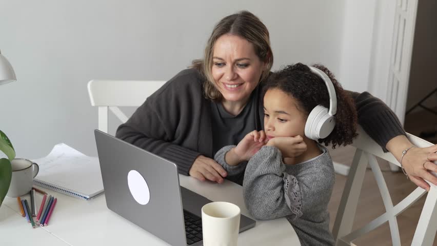 Caucasian mother and African American daughter studying at home, mom teaching doing homework in cosy white room using laptop. Parent online education, child school girl learn, type test exercise