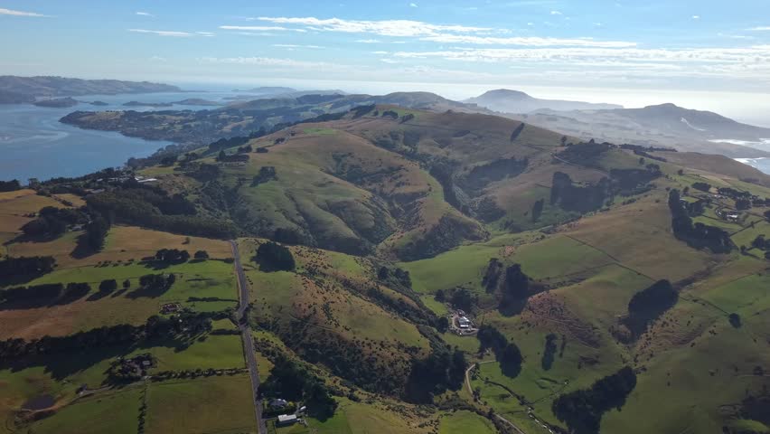 Sideways drone flight over lush green fields, prairies, houses, and the distant ocean, revealing the peaceful and scenic charm of Dunedin, New Zealand.