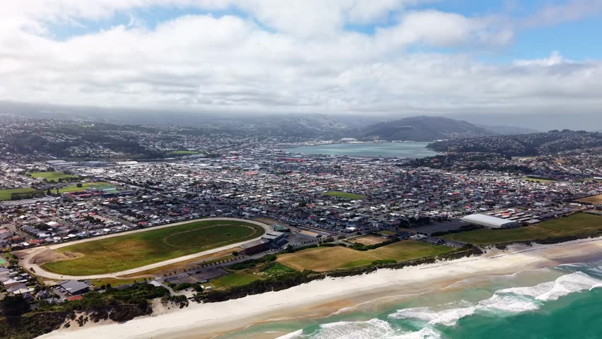 Drone shot pulling back and ascending above Dunedin, showing the coastline, sports field, harbor, and cityscape under cloudy skies