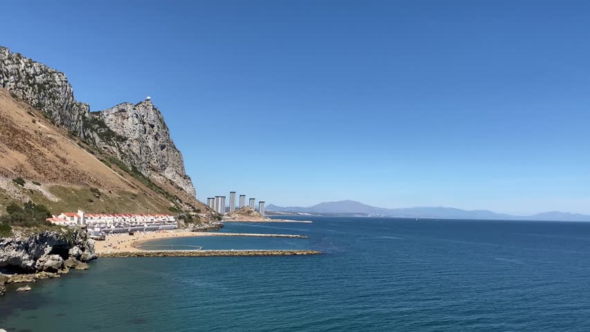 Wide angle view of the eastern side of Gibraltar with Sandy Bay and Catalan Bay resorts in the background. The calm Mediterranean sea basks under a sunny clear blue sky.