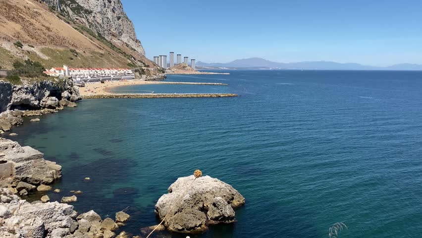 Wide angle view of the eastern side of Gibraltar with Sandy Bay and Catalan Bay resorts in the background. The calm Mediterranean sea basks under a sunny clear blue sky.