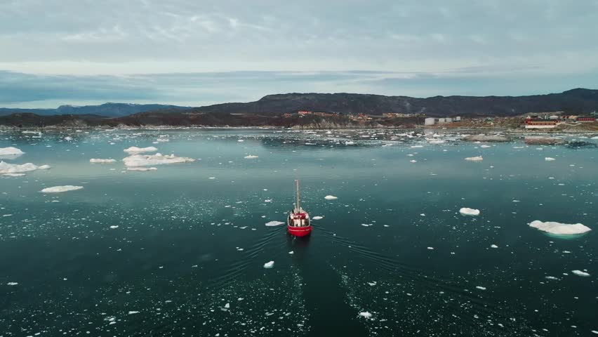 A red fishing boat sails through icy Arctic waters near Greenland, surrounded by floating icebergs and snow-capped mountains in the distance, captured by a drone on a calm, cold day.