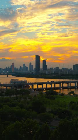 Vertical aerial wide angle view of Banpo Bridge and expressway traffic over Han River, Hangang Park greenery at sunset, with Raemian Caelitus Tower 101 and 63 Building lighting up Seoul's skyline