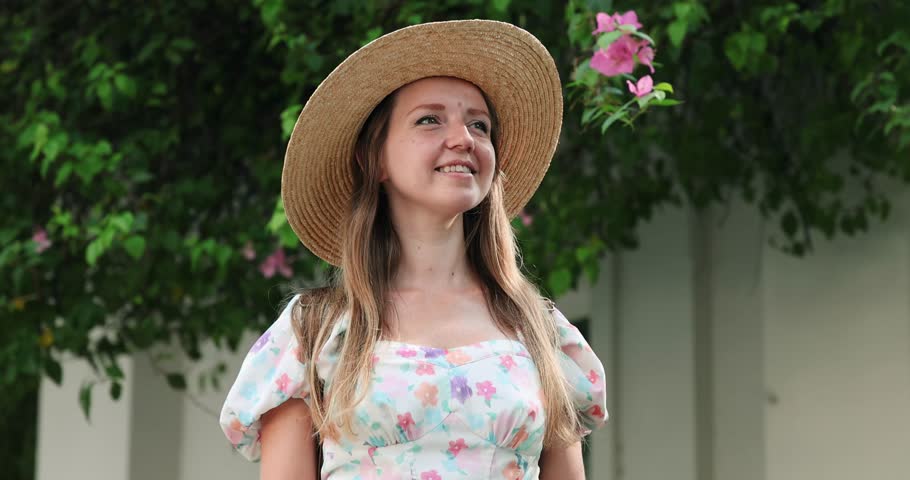 Woman in floral dress with straw hat, smiling outdoors near blooming garden, capturing essence of spring and nature.