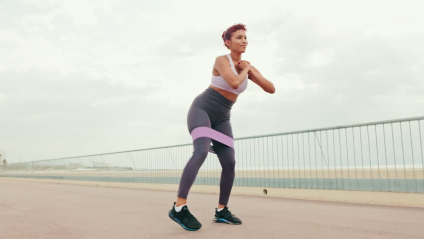 A young woman performs squats and leg raises with a resistance band. She is exercising outdoors on a paved path next to a railing and beach.