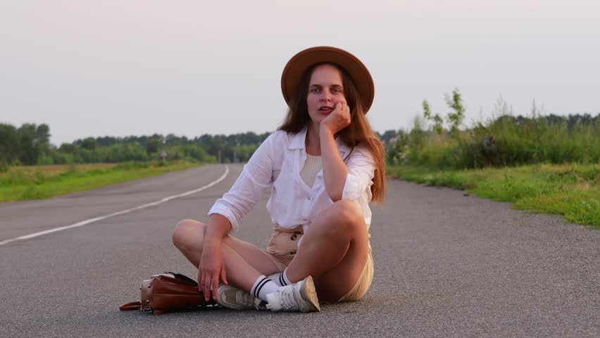 A young woman sits cross-legged on a quiet country road during sunset, wearing a hat and looking contemplative while surrounded by nature