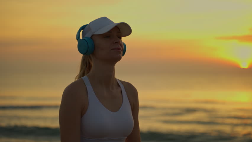 Silhouette of joyful woman wearing headphones and stretching on beach at sunrise, immersing herself in music, yoga, and tranquil morning routine by sea