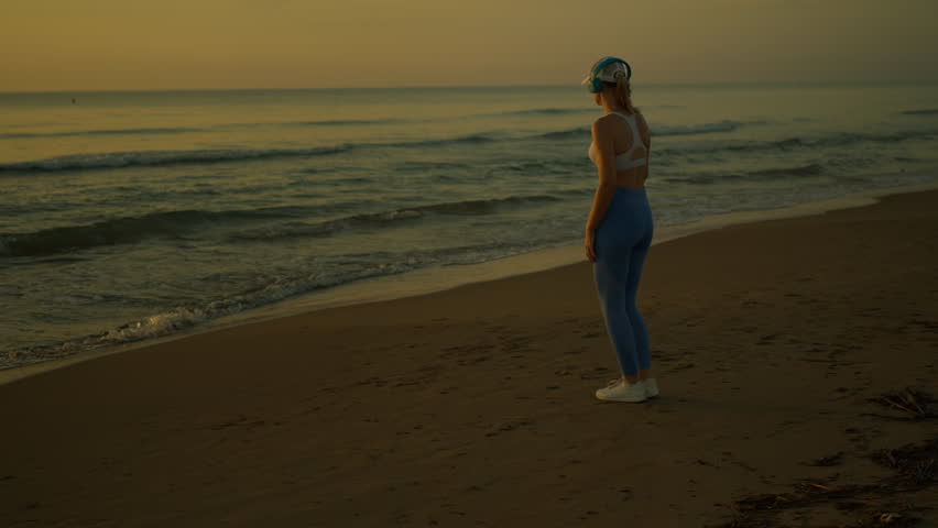 Beautiful athletic woman in sportswear warming up and stretching on sandy beach at sunrise while listening to music and waves