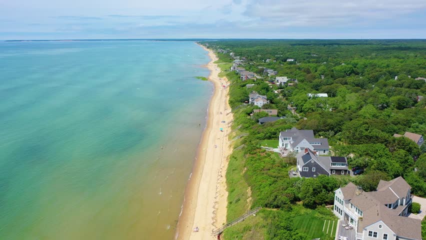 Maine beach scene with families wading in turquoise water, kayaks drifting nearby, and traditional coastal cottages dotting the shoreline framed by lush green trees and open sandy beaches.