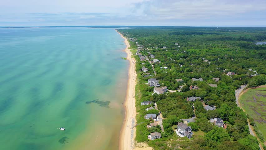 Drone shot reveals a wide sandy shoreline where families sunbathe under umbrellas, swimmers wade in shallow turquoise waters, and boats rest offshore beyond coastal homes and tree cover.
