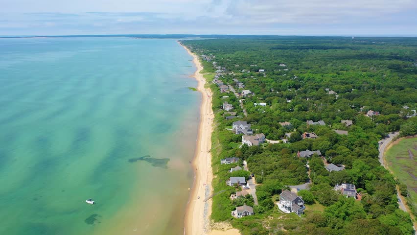 People sunbathe on golden sand while children wade in shallow turquoise water, kayakers paddle offshore, and charming beach houses overlook the lively coastal scene on a warm summer day.