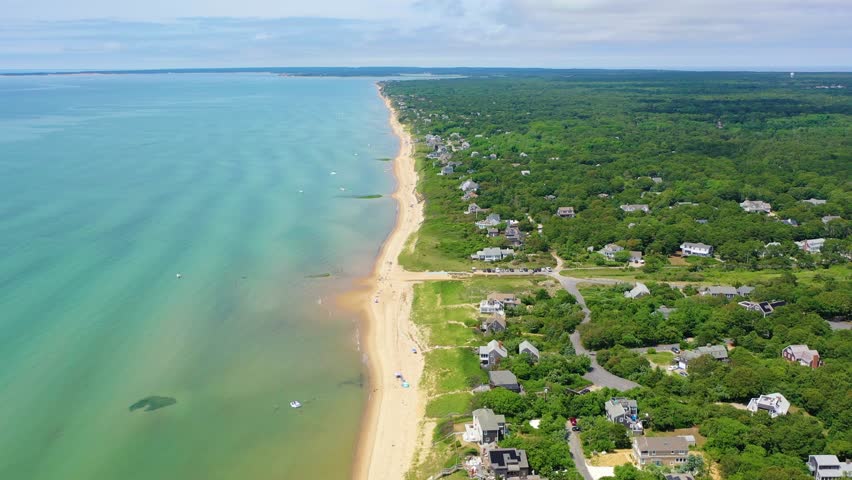 Drone video captures Cape Cod shoreline with swimmers wading, paddleboarders gliding offshore, and classic beach houses perched on bluffs surrounded by green trees above the sandy shore.