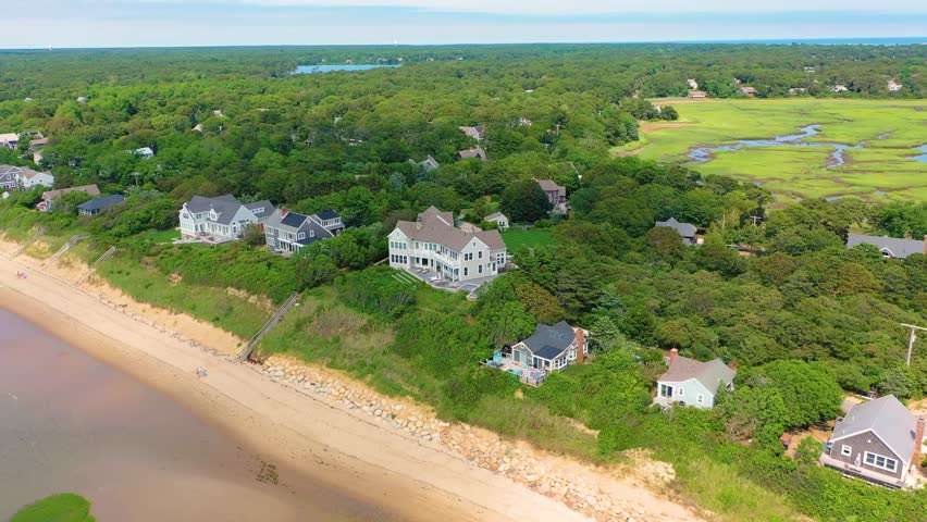 Aerial orbit of Cape Cod captures a beach house above sandy dunes, turquoise water reflecting sunlight across the coast, and families strolling nearby.