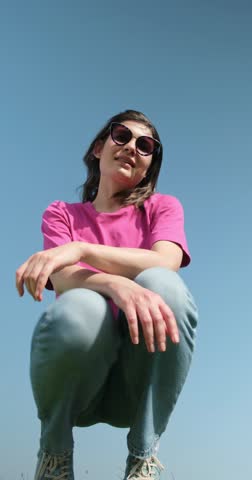 Vertical video of beautiful carefree young woman in sunglasses with curly hair in pink t-shirt and blue jeans sitting and looking down at camera on blue sky background low angle shot.