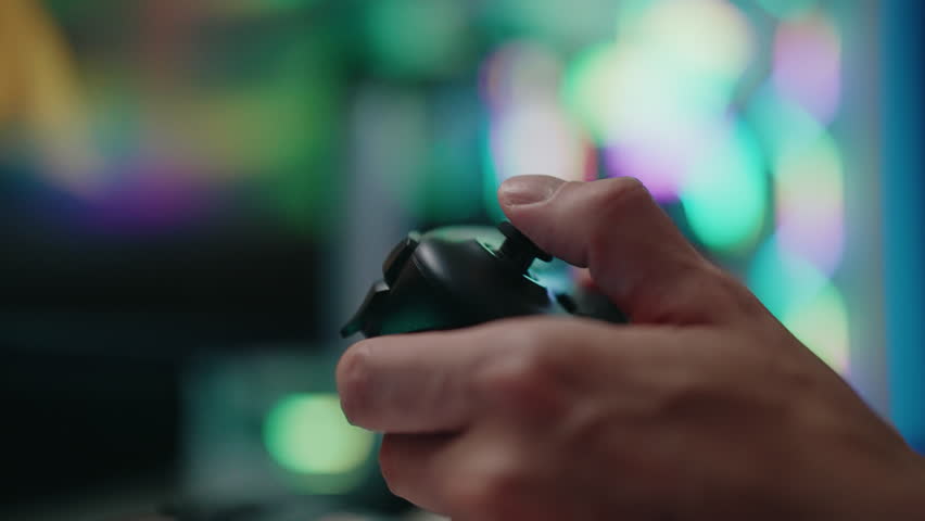Gamer holding using wireless black controller, engaging with vibrant video game in dark room illuminated by neon lights from computer case on blurred background, creating immersive gaming experience