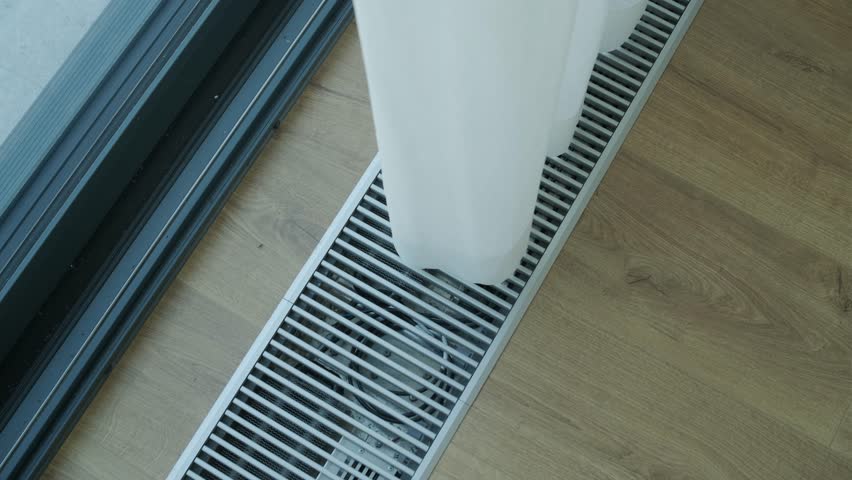 Metal floor vent grille near a large window on wooden flooring, part of a modern interior ventilation or heating system.