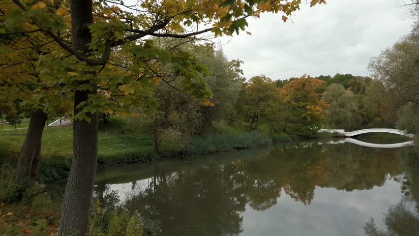 Peaceful autumn scenery with lush deciduous trees covered by colorful leaves on shore of city lake or pond with arch bridge at cloudy october dusk. No people nature scenic autumnal landscape.