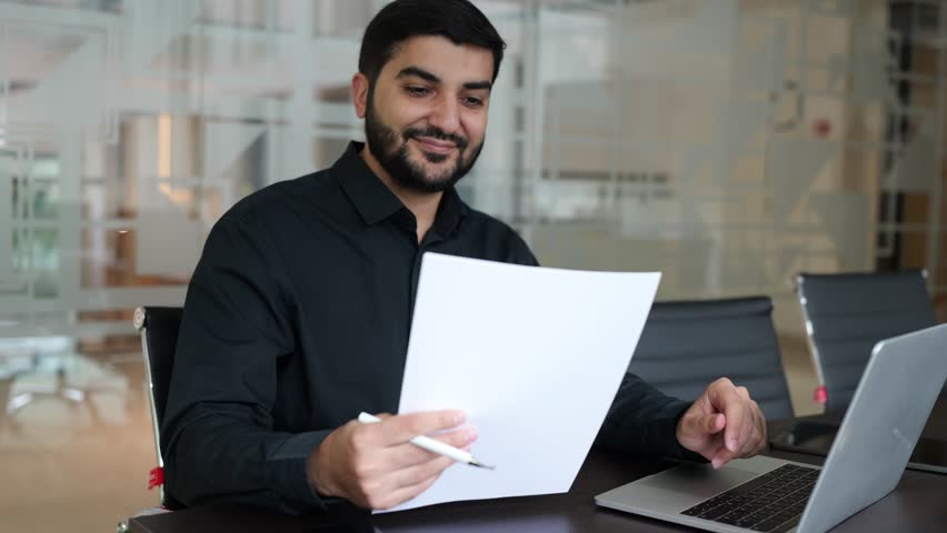 Male professional is seated at a desk in a contemporary office, examining important documents while a laptop and phone are nearby, deeply engaged in his task.	
