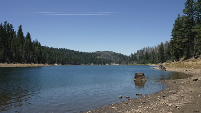 Lake with shoreline and conifer forest against mountain backdrop, creating an atmosphere of tranquility and natural beauty on a clear sunny day