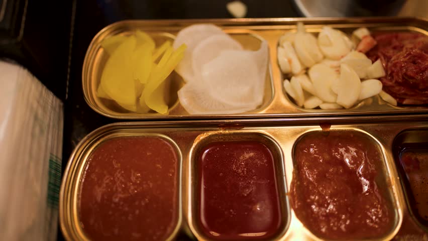 Overhead camera rotates above metal tray with Korean BBQ banchan, garlic, kimchi, dipping sauces