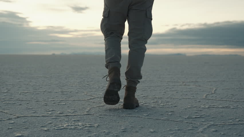 Close up of a man’s boots walking across the cracked white surface of the Uyuni salt flats in Bolivia during a cloudy morning in slow motion