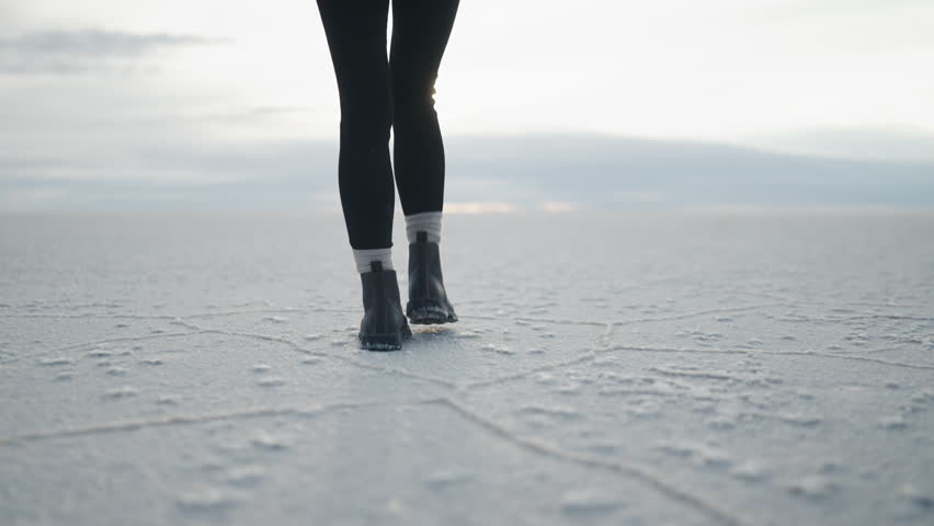Close up of a woman’s boots walking across the cracked white surface of the Uyuni salt flats in Bolivia during a cloudy morning in slow motion