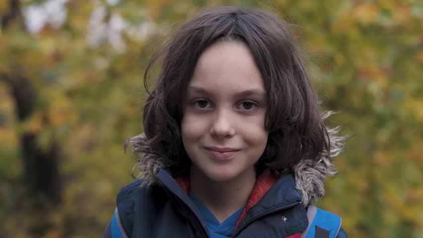 A cute boy with long hair puts on the hood of his jacket while standing on the street. A child on the street.
