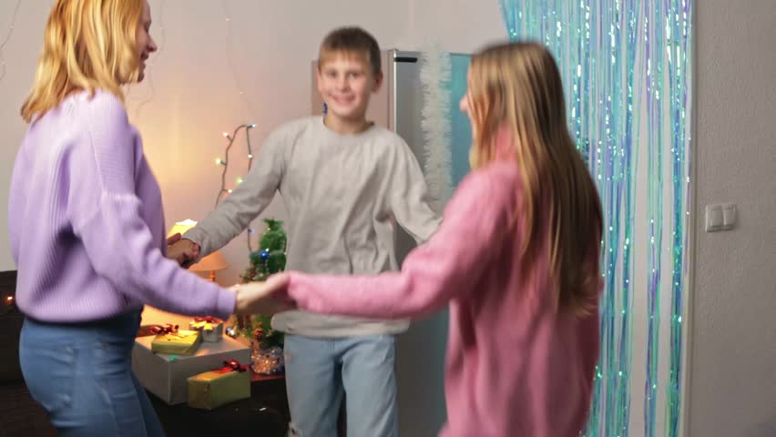 Happy mother with her daughter and son dancing in circle holding hands in decorated room, enjoy family party and celebrating christmas holiday with festive lights