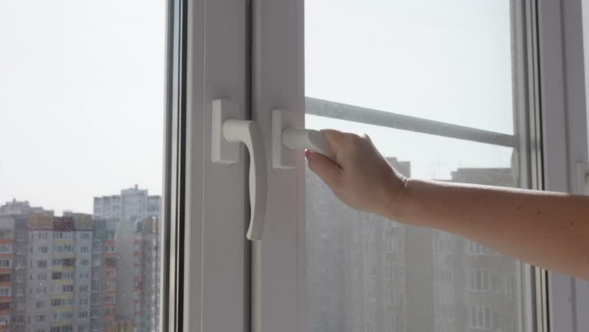 Female hand turning white window handle and opening modern plastic window frame revealing city buildings and cloudy sky, fresh air and ventilation in residential apartment.