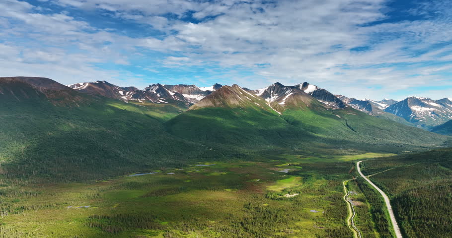 Shadows of clouds covering the stunning snow-capped mountains. Highway crosses the pine tree wood in the valley. Alaska, USA.