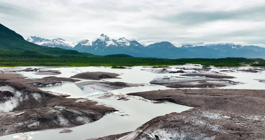 Footage over the melting ice and dirty waterscape. Beautiful green waterfront and snow-capped mountains of Alaska at backdrop.