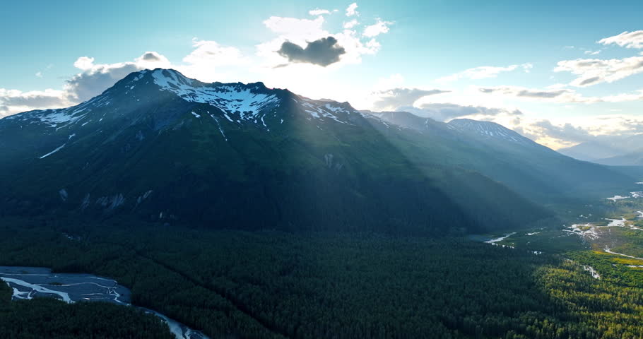 Little cloud covers the sun over the stunning mountain. Rays of sun light the valley covered with pine tree woods. Alaska, USA.