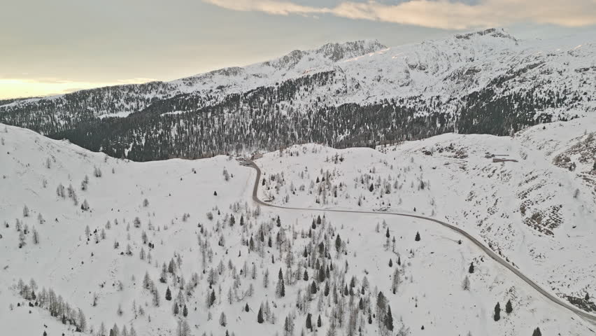 Top-down style drone shot of a serpentine road cutting across snowy hills dotted with conifers and cabins at dusk.