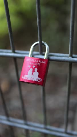Vertical screen red love padlock attached to metal fence symbolizing eternal affection and romantic promise between couple shining in soft daylight creating intimate emotional atmosphere of devotion