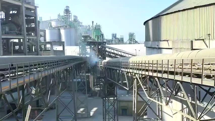 A wide-angle industrial view of a cement factory with multiple storage silos and a network of conveyor belts transporting raw materials.
The footage shows the intense process of material transport, 