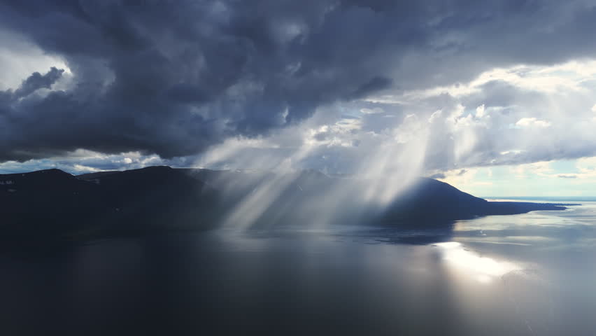 Aerial view of brooding ridges and wide water as sunray pierce the cloud bank. Blue haze drifts across the valley, revealing untouched taiga, long shoreline and a tourism gem in the heart of Siberia