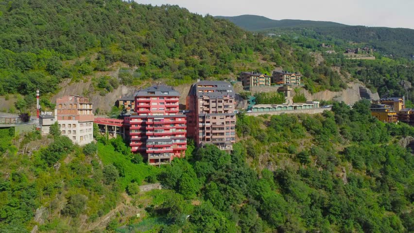 Drone footage captures a stunning aerial perspective of modern residential buildings nestled amidst the verdant, tree covered mountains of Andorra. The vibrant architecture stands out against the natu