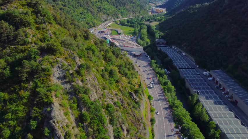 An expansive aerial shot showcases the stunning natural beauty of Andorra, featuring winding mountain roads, verdant valleys, and dense green forests nestled within the Pyrenees.