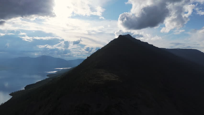 Evening drone flyover reveals a sharp volcanic ridge casting a long shadow over Lake Lama, sunbeams breaking through heavy cloud and glitter on the water. Slow forward motion builds scale and a sense