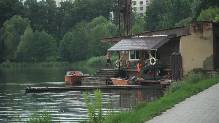 A man with athletic build black shorts jumps rope terrace house lake, small boats are moored nearby, old fishing house, lake city center, tall green trees around, cloudy summer day