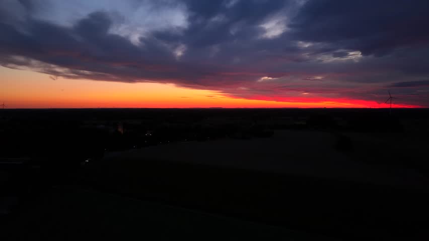 Dark landscape and suburb town in American during bright colorful sunrise at horizon. Rotating wind turbine silhouette. Aerial rising wide shot.