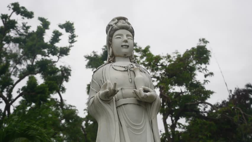 A low static angle holds throughout, framing a towering statue in serene stillness. Robed and adorned, the figure stands poised in symbolic gesture, surrounded by trees and clouded sky.
